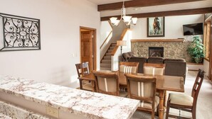 A dining area with a marble countertop and wooden chairs, a staircase leading to a living room with a stone fireplace, and a mounted artwork on the wall.