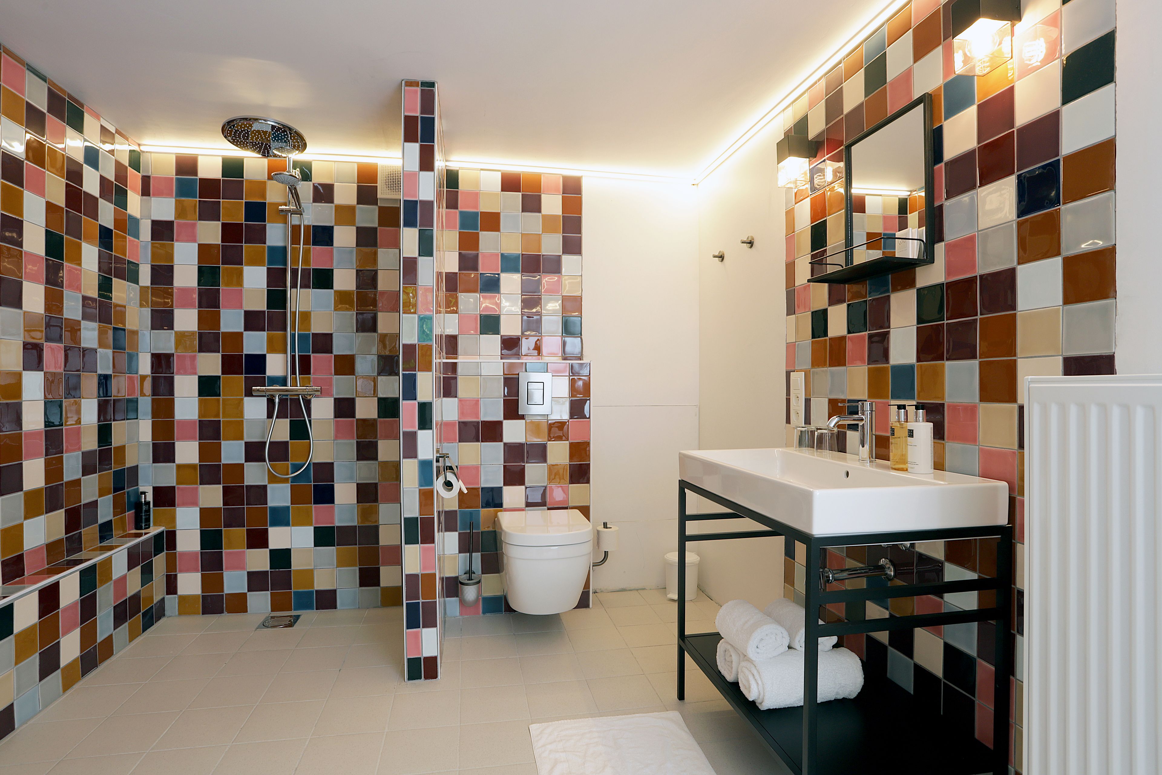 Un baño con pared de ducha enlosetada, inodoro, lavabo con espejo y estante con toallas.