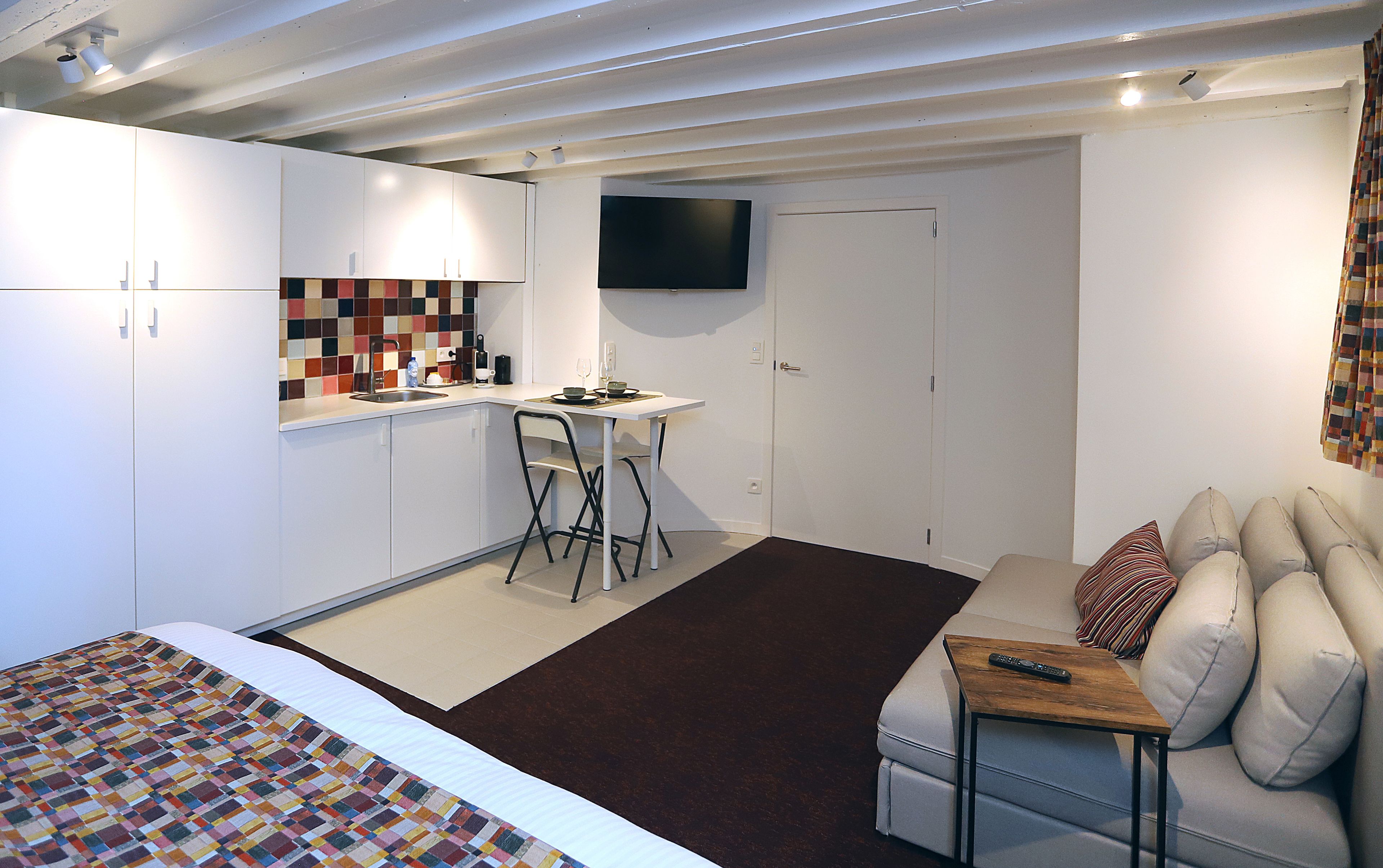A compact hotel room with a kitchenette, a dining area, a TV mounted on the wall, and a sofa with a remote control on a small table.