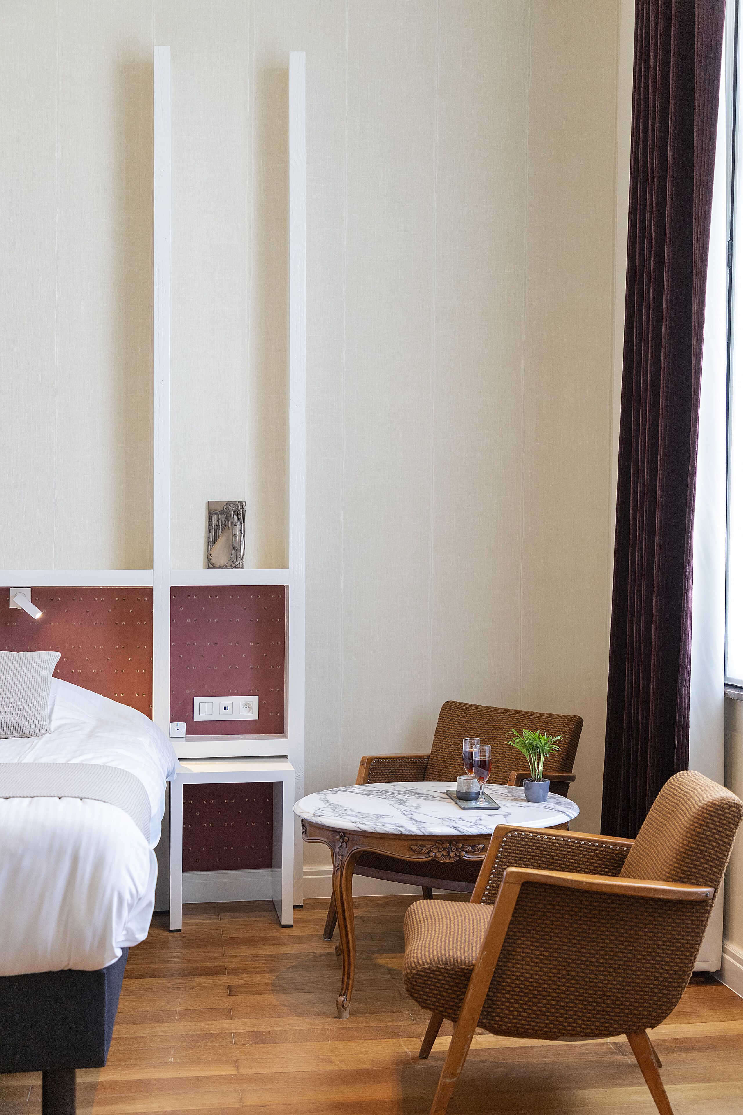 A hotel room with a bed, a small table with a wine glass and a plant, two wicker chairs, and a wall-mounted shelf with a picture.