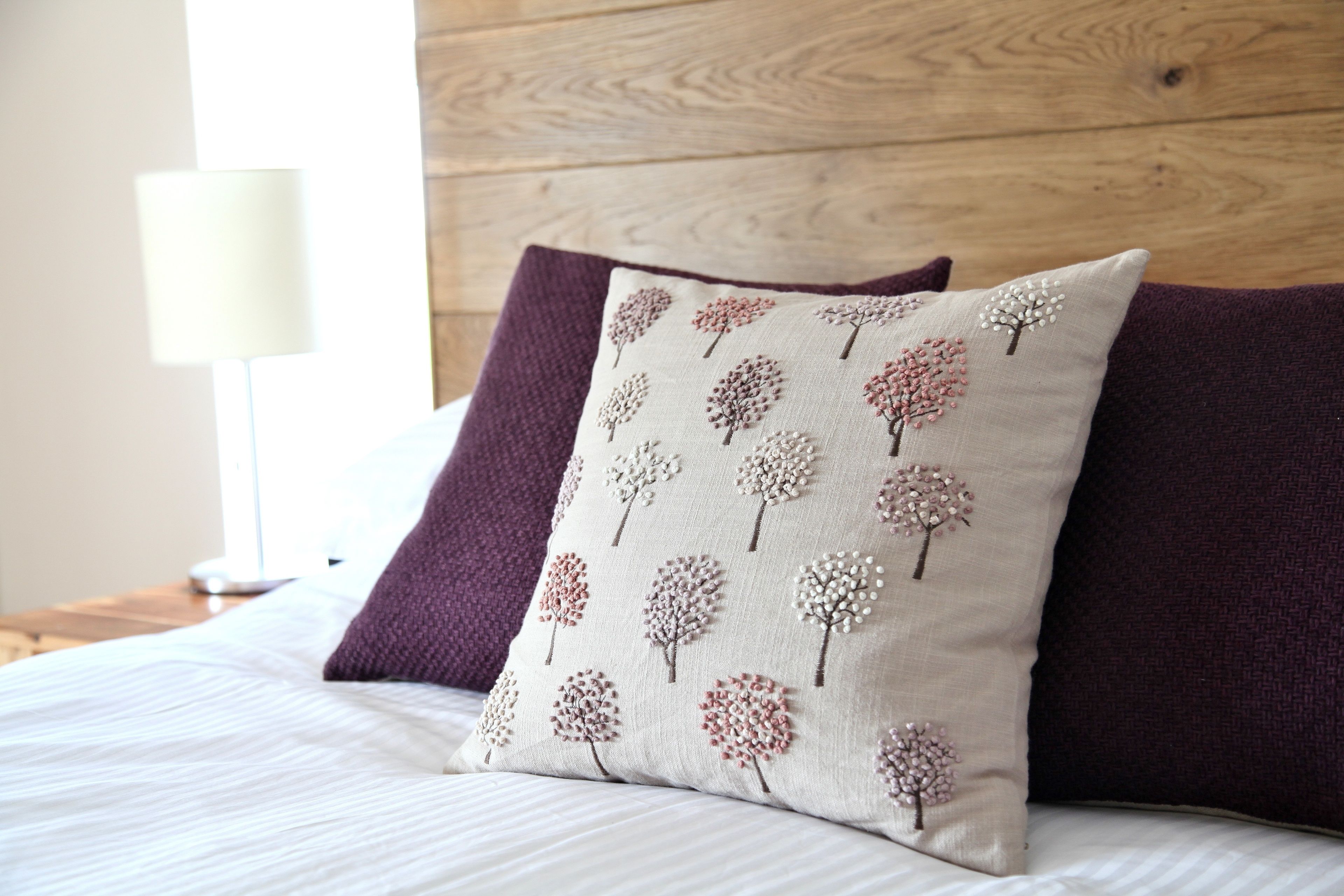 Un lit avec un oreiller beige orné d’un motif d’arbre, un oreiller violet et une lampe blanche en arrière-plan.