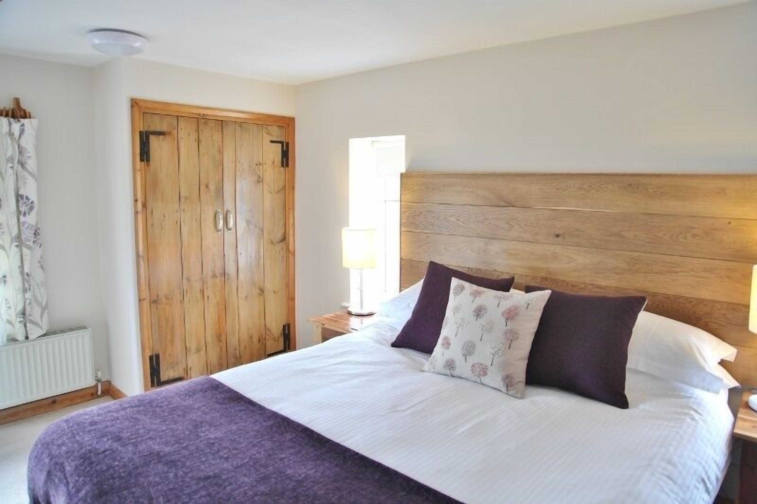 Een slaapkamer met een houten hoofdbord, een bed met wit en paars beddengoed, een nachtkastje met een lamp en een houten kledingkast.