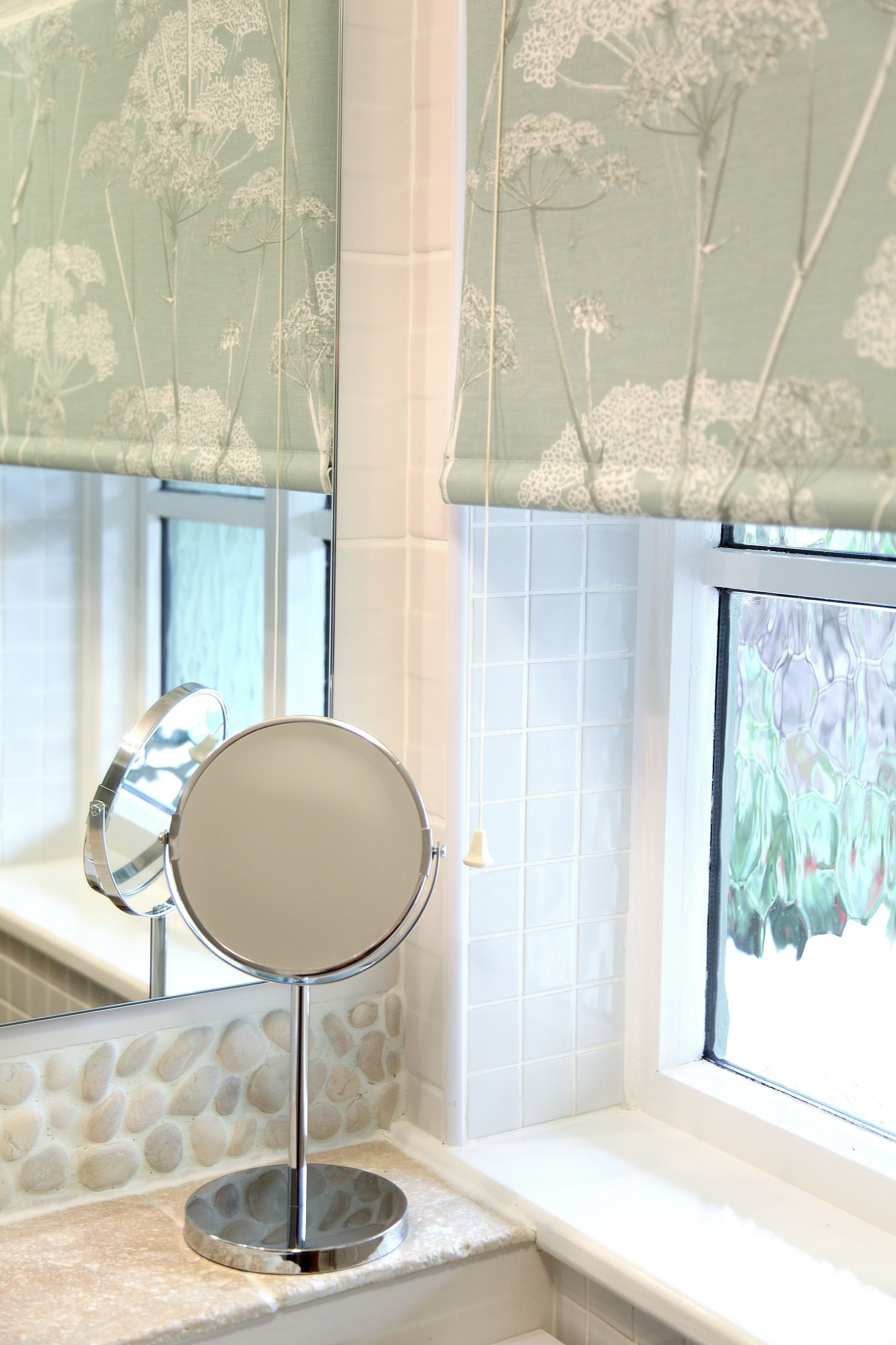 Un baño con un espejo redondo sobre un pedestal, una ventana con una persiana floral y una pared con diseño de piedra.