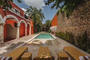 Un espace piscine avec des chaises longues, un mur en pierre et un bâtiment rouge orné d’arches.