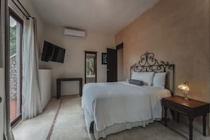 Un dormitorio con cama, mesita de noche, lámpara, televisor montado en la pared y ventana con cortinas.