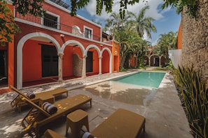 A poolside area with lounge chairs and a swimming pool, surrounded by a red building with arches and a stone wall.