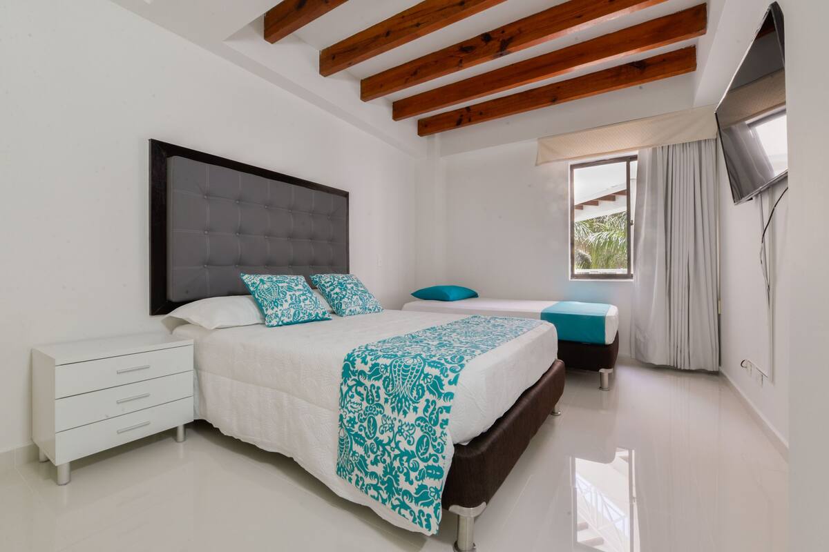 Un dormitorio con cama, mesitas de noche, televisor y ventana con cortinas.