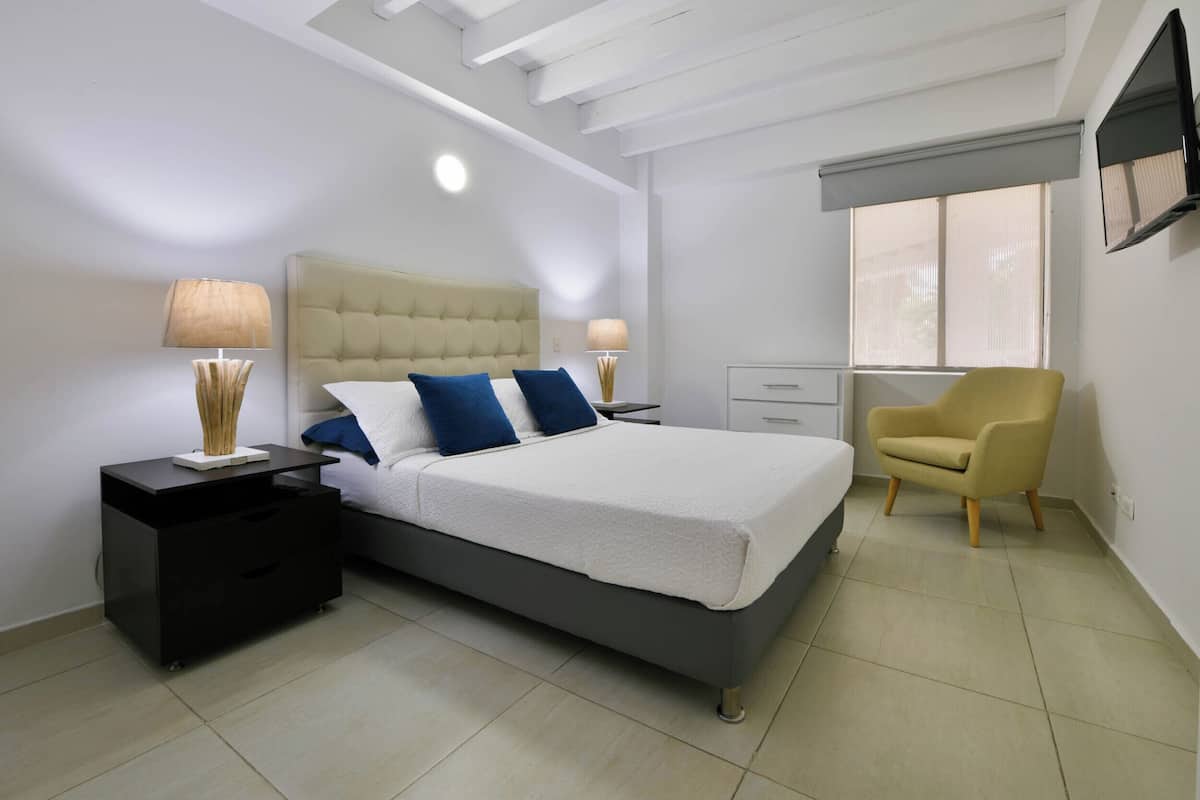 Un dormitorio con cama, mesitas de noche, una silla, televisión y ventana.