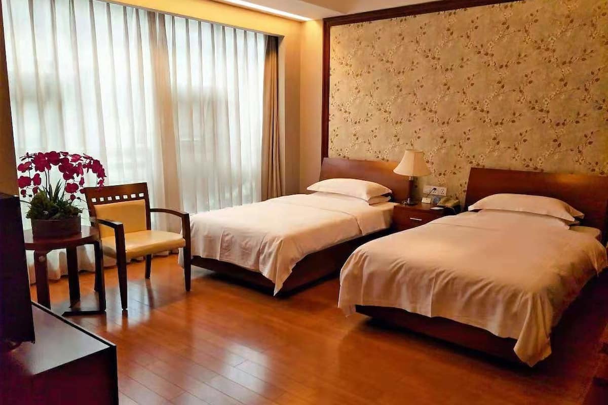 Habitación de hotel con dos camas, piso de madera, escritorio con silla, planta en maceta y ventana con cortinas.