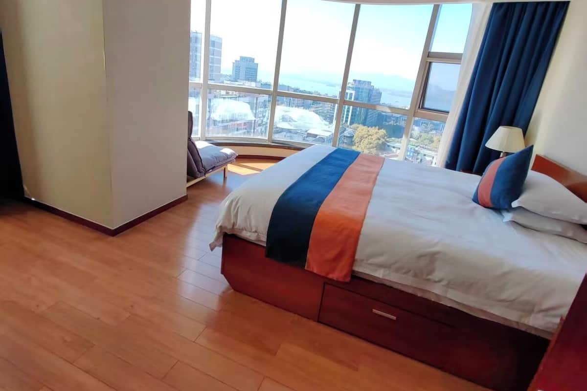 Habitación de hotel moderna con una cama grande, vista a la ciudad a través de amplios ventanales y un televisor de pantalla plana en el techo.