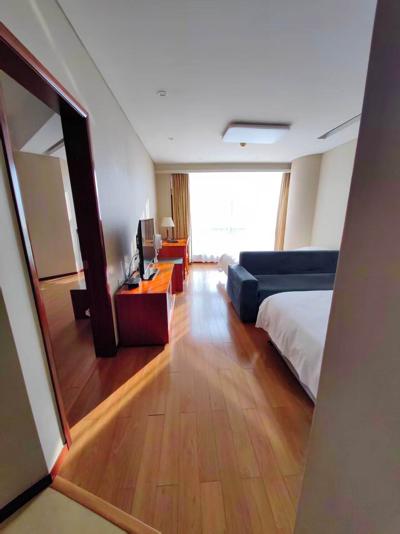 Una habitación de hotel con piso de madera, una cama, un escritorio con computadora, una silla y una ventana con cortinas.