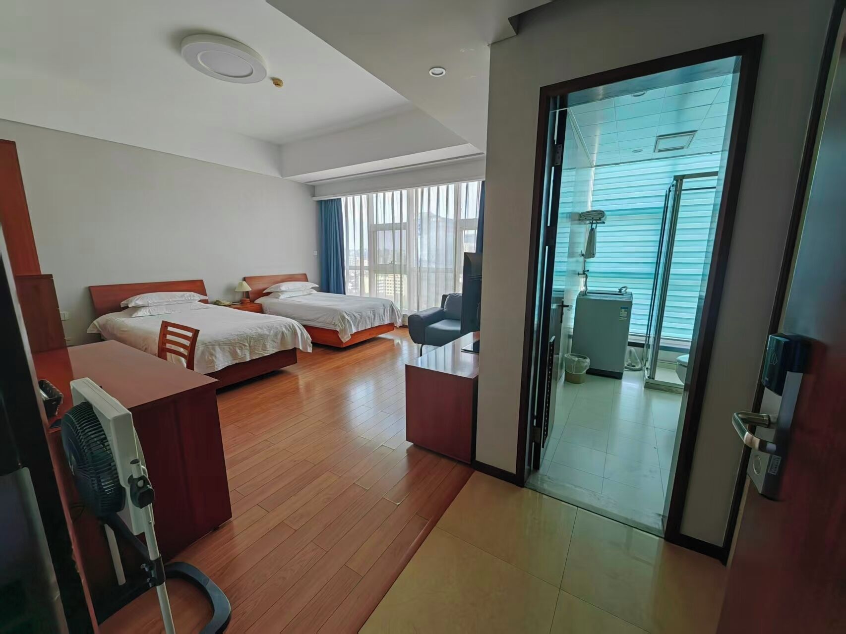 A hotel room with two beds, a desk, a chair, a fan, and a sliding glass door leading to a balcony.