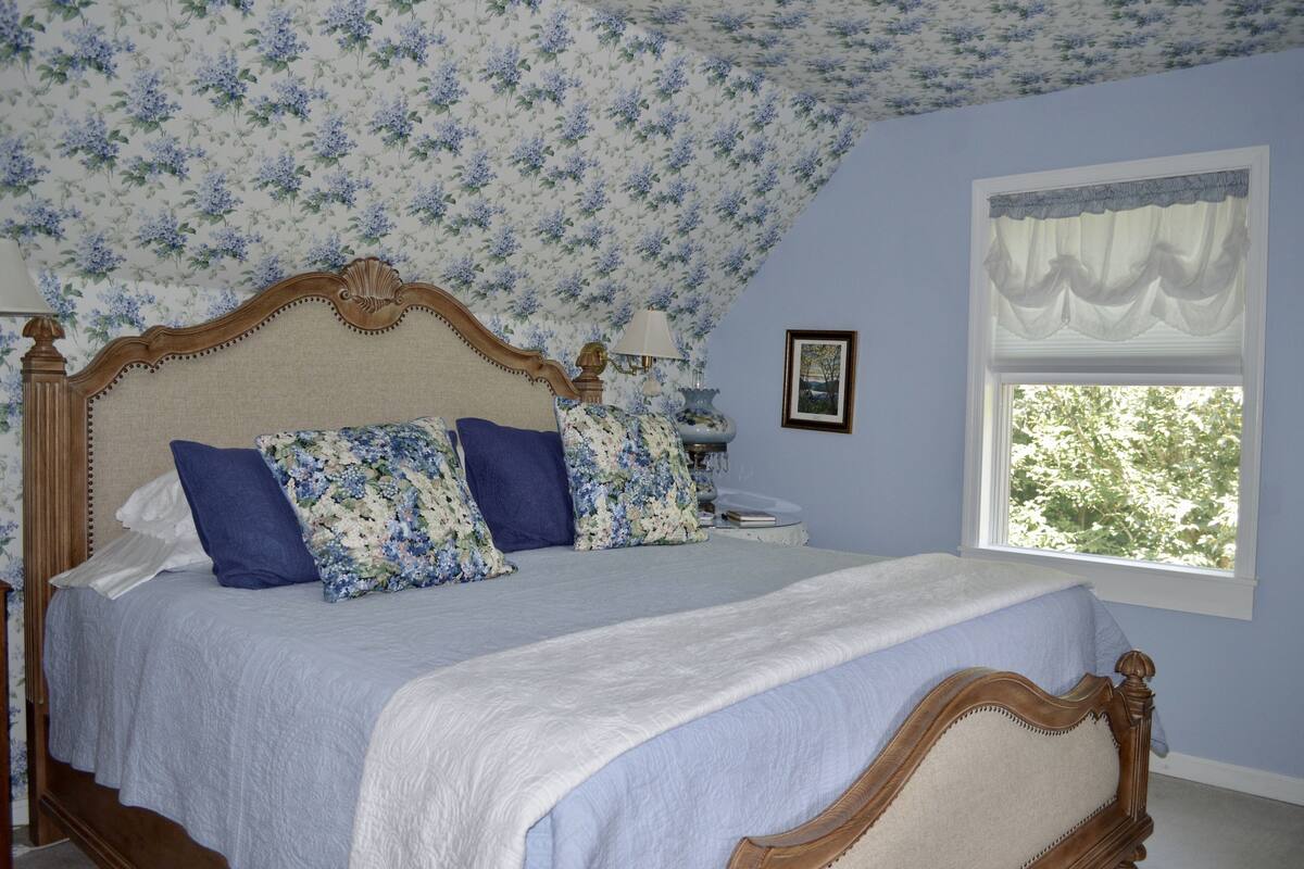 A bedroom with a floral-patterned ceiling, a large bed with a wooden headboard, and a window with a white valance.