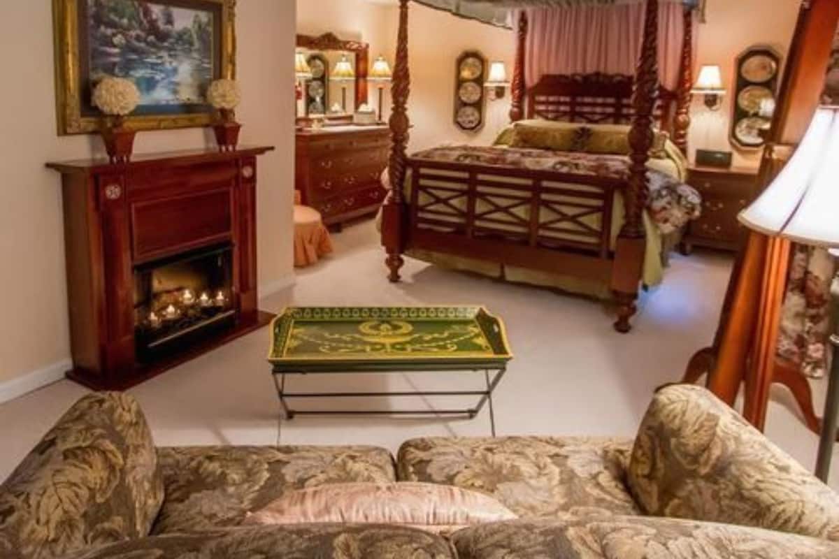 A traditional bedroom with a four-poster bed, a fireplace, and a green ottoman.
