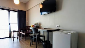 Un espacio habitable compacto con kitchenette, televisor de pantalla plana montado en la pared, comedor con mesa y sillas, y puerta de vidrio corrediza que da a un balcón.