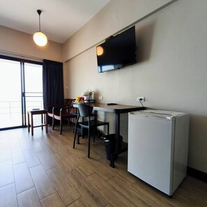 Un espace de vie compact comprenant un coin cuisine, un téléviseur à écran plat fixé au mur, un coin repas avec une table et des chaises, et une porte-fenêtre coulissante donnant sur un balcon.