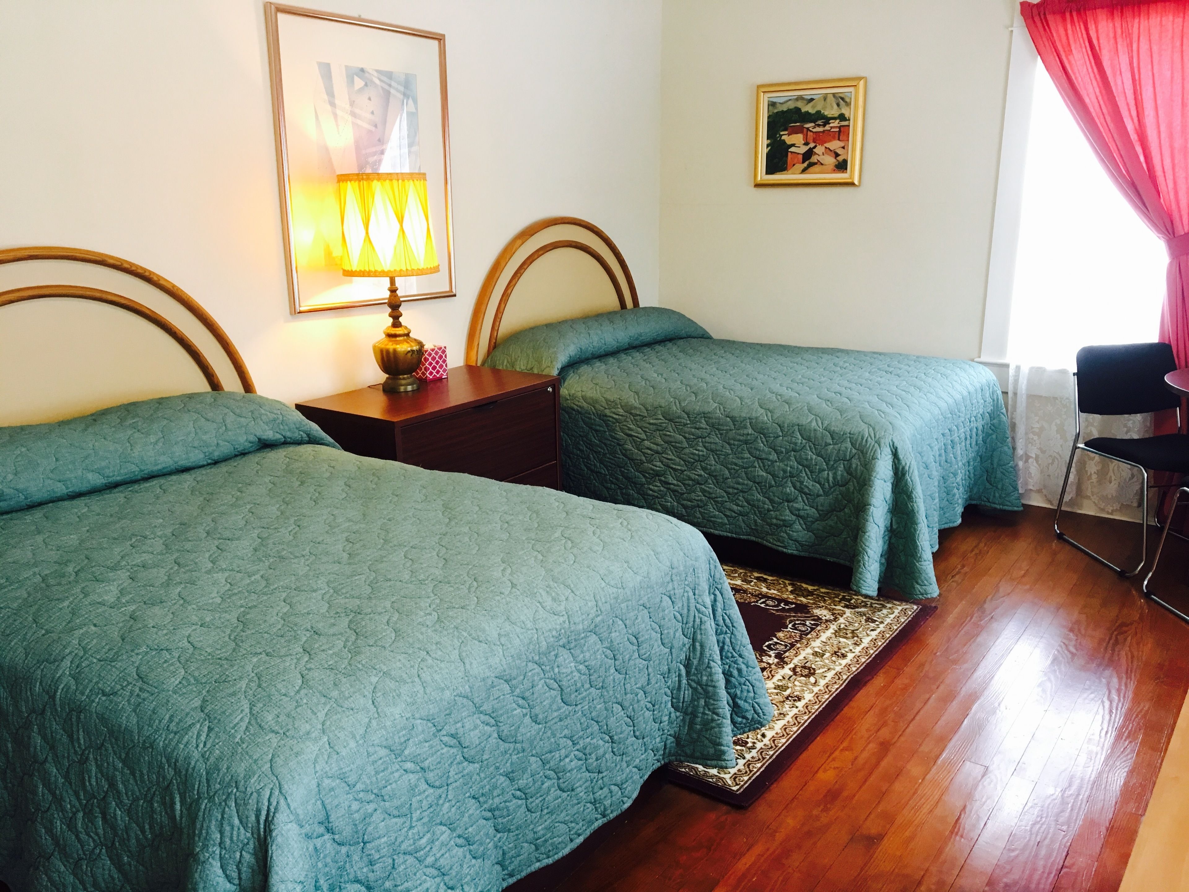 Habitación de hotel con dos camas individuales, una mesita de noche de madera con lámpara, una pequeña mesa, una silla y una alfombra.