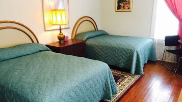 Habitación de hotel con dos camas individuales, una mesita de noche de madera con lámpara, una pequeña mesa, una silla y una alfombra.