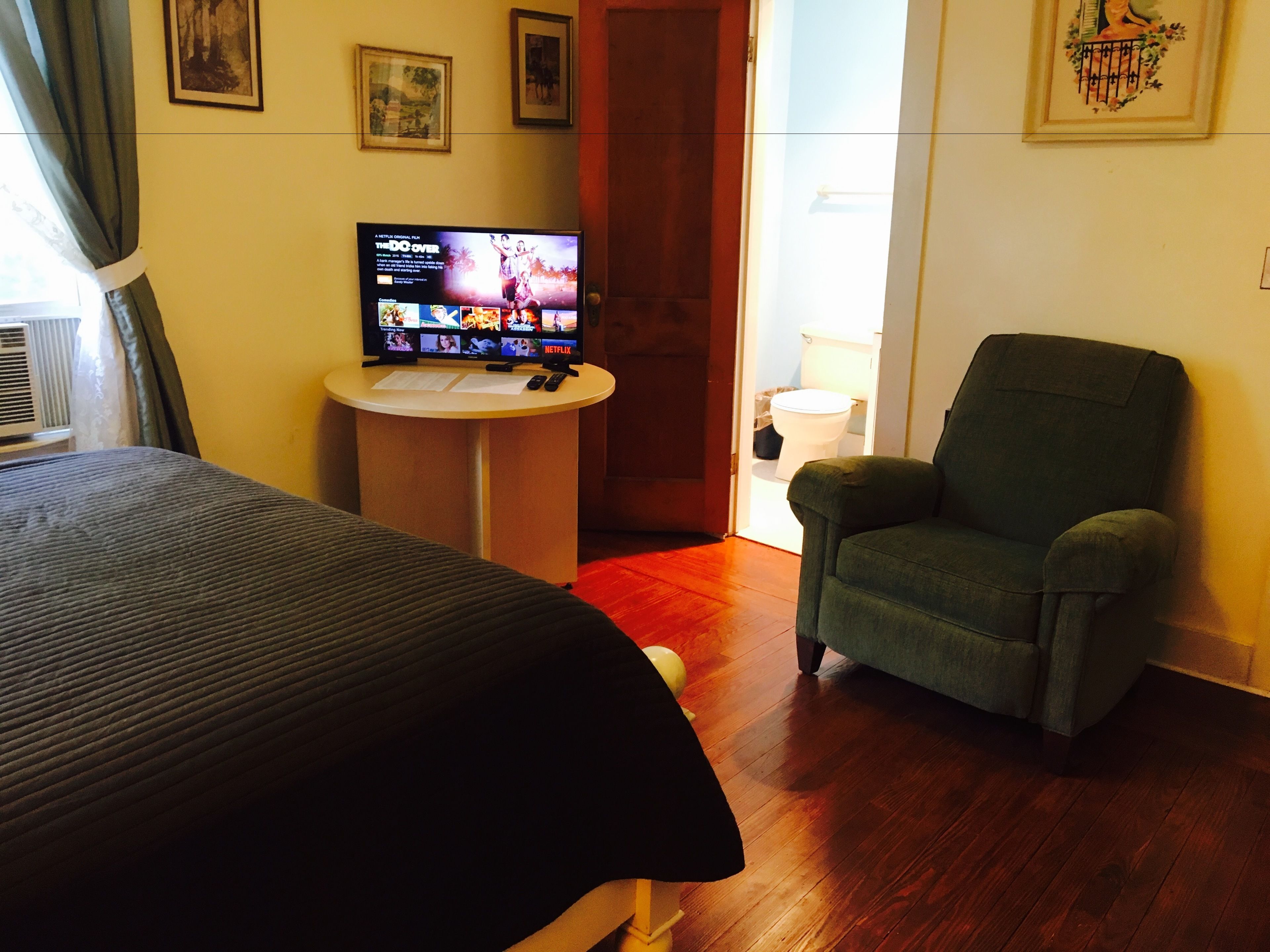 Habitación de hotel con cama, un sillón verde, una mesita con un televisor mostrando una película y una puerta que da a un baño.