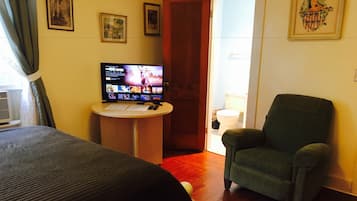 Habitación de hotel con cama, un sillón verde, una mesita con un televisor mostrando una película y una puerta que da a un baño.