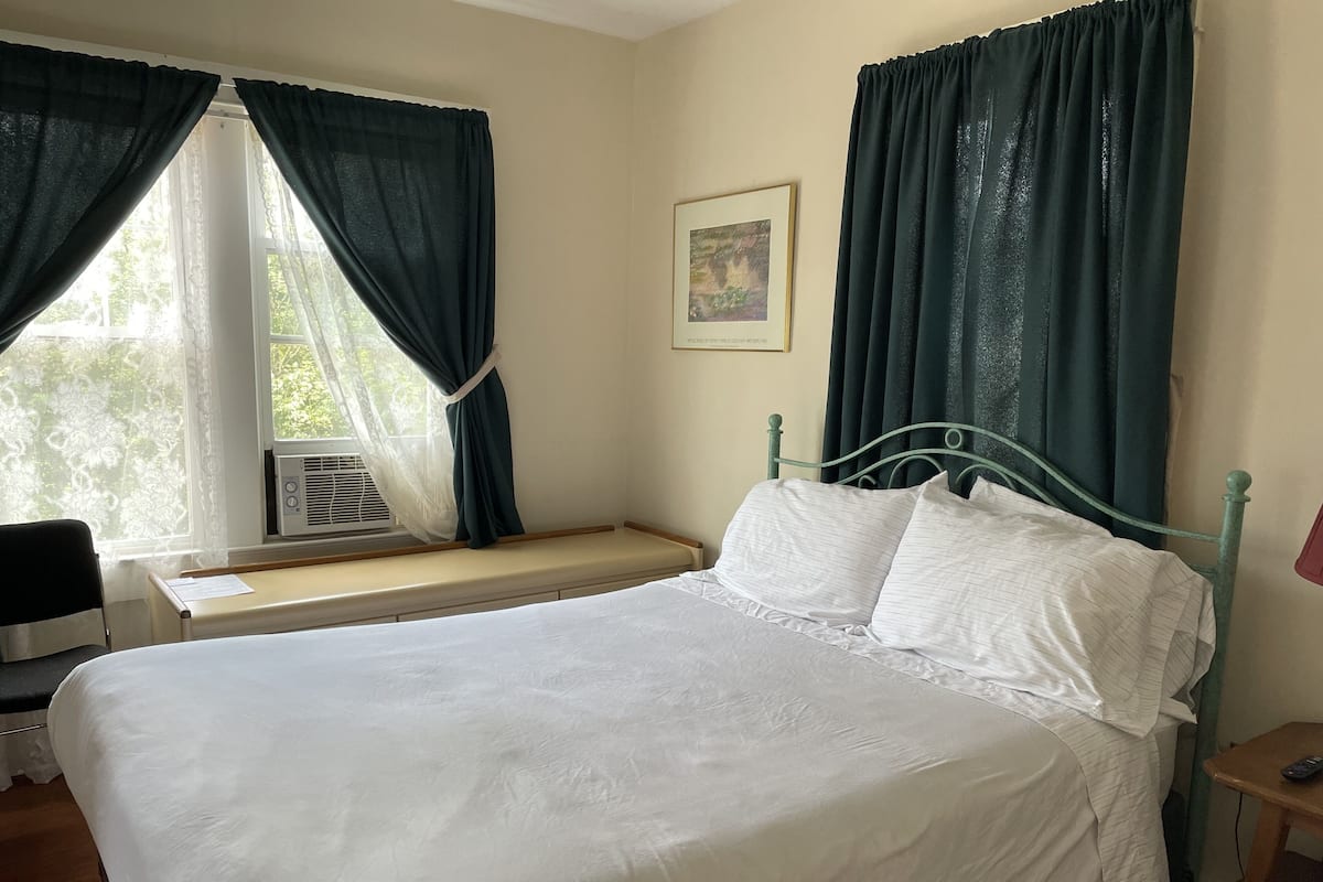 Un lit bien fait, avec du linge de lit blanc, une fenêtre ornée de rideaux légers, une table de chevet avec une lampe et un tableau encadré accroché au mur.