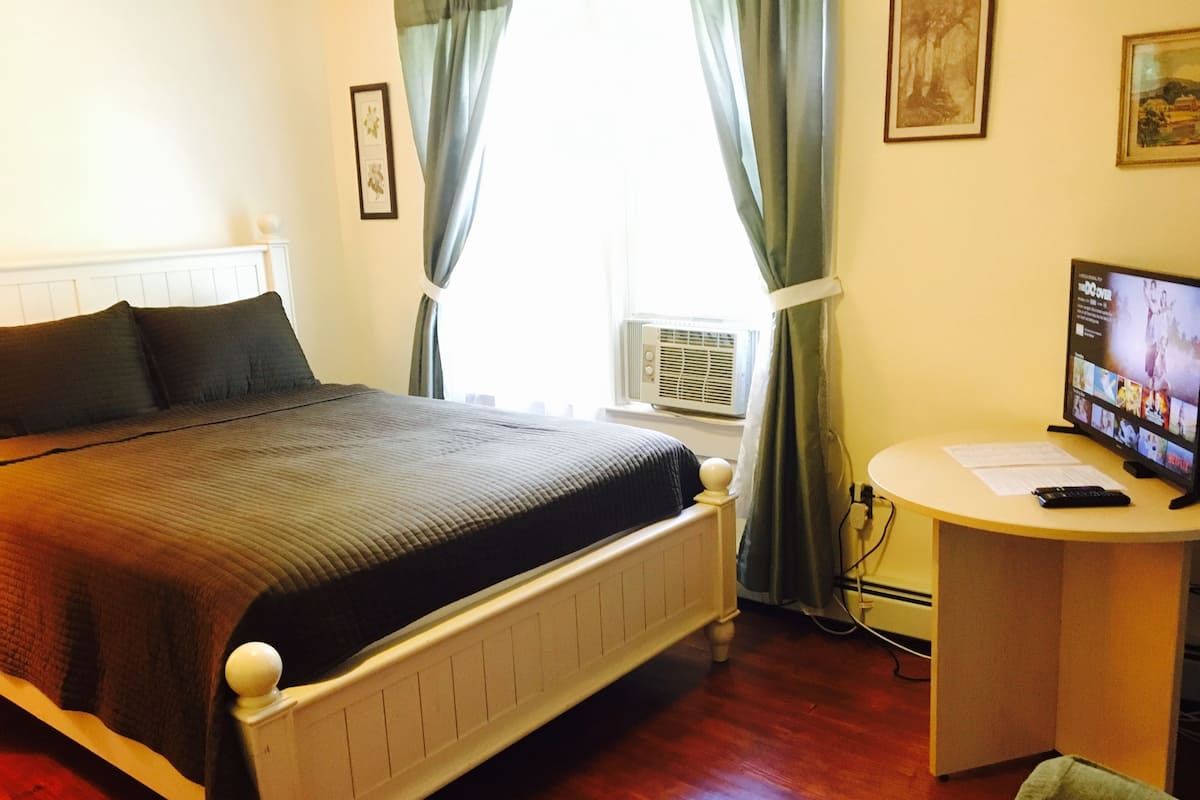 Une chambre à coucher avec un lit, un bureau avec un ordinateur et une fenêtre avec des rideaux.