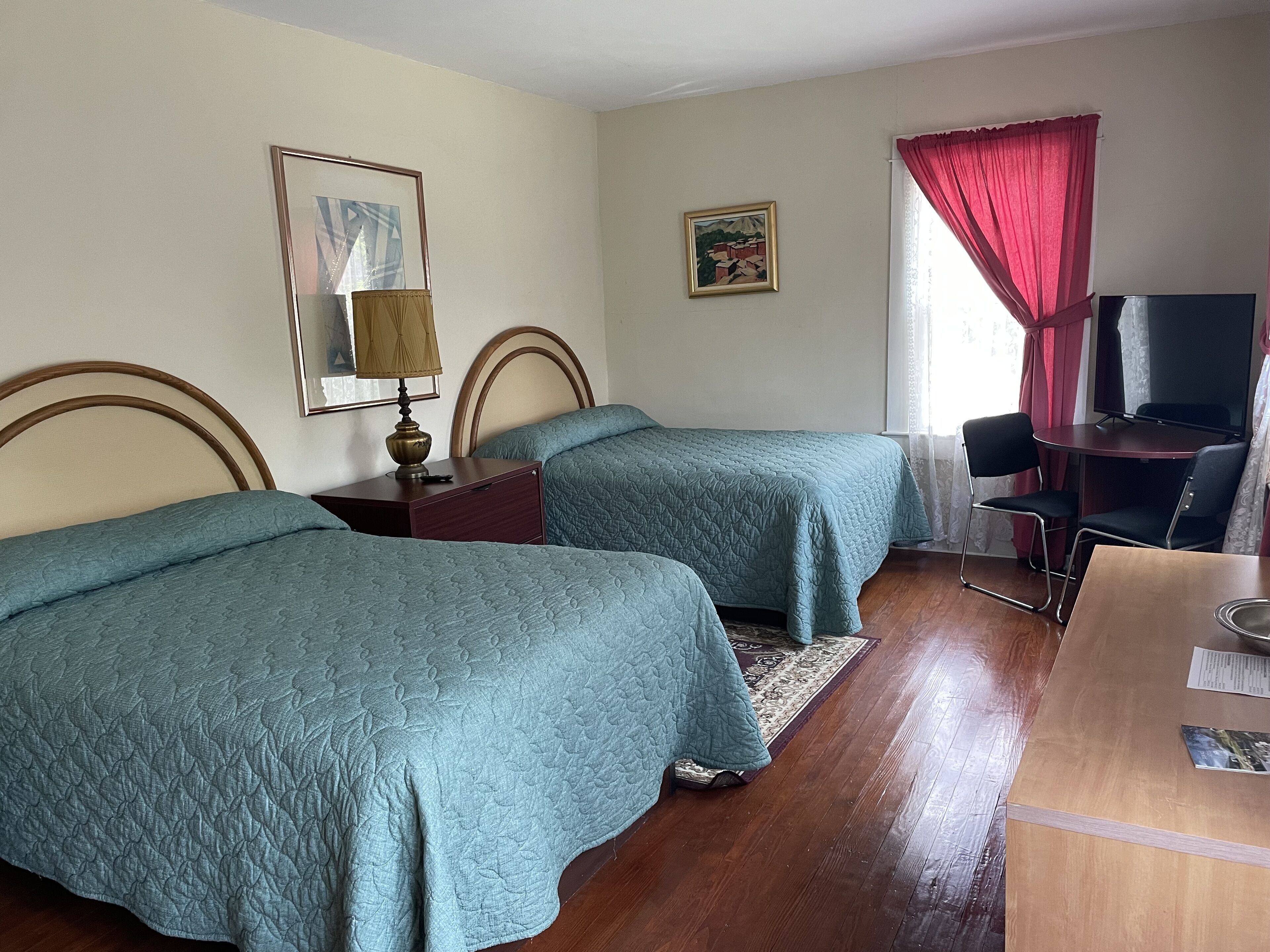 A hotel room with two single beds, a desk with a chair, a TV, and a window with red curtains.