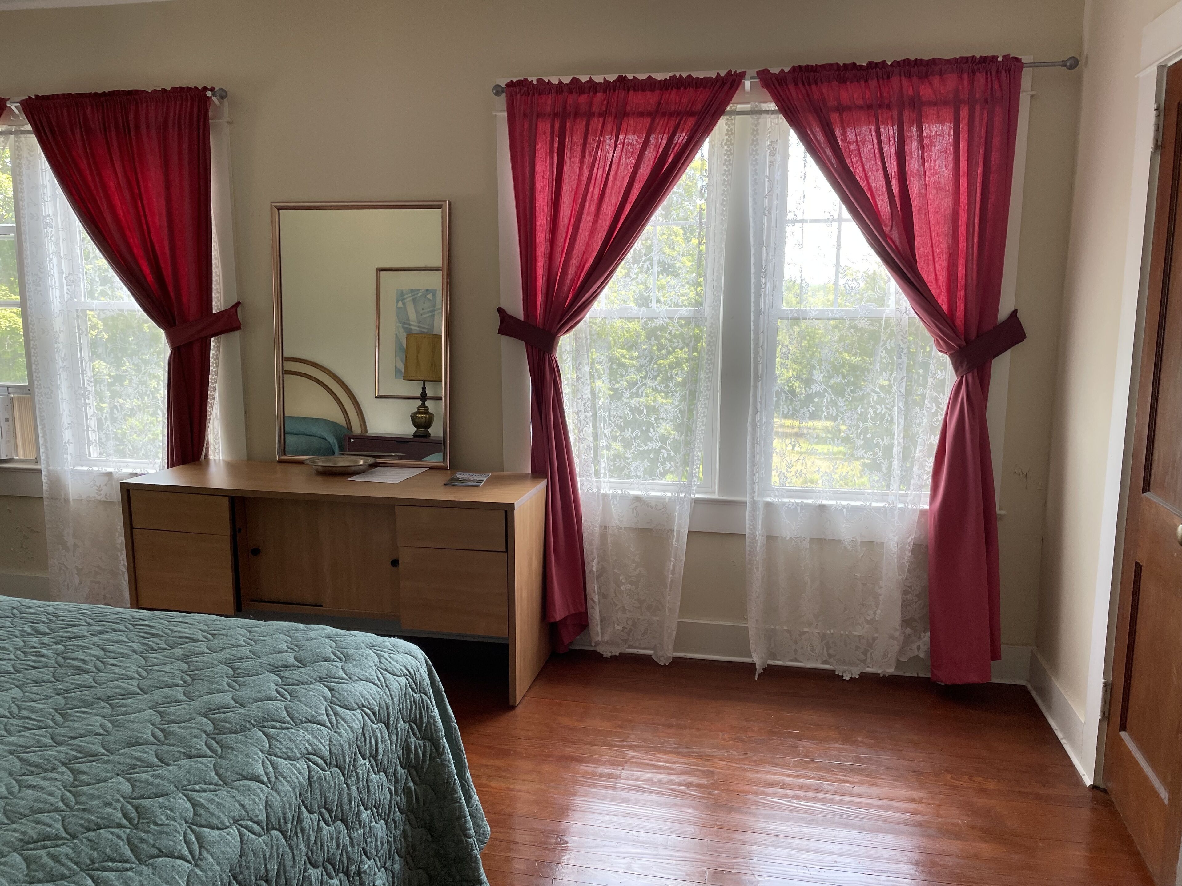 A bedroom with a wooden dresser, a bed with a green quilt, two windows with red curtains, and a mirror above the dresser.