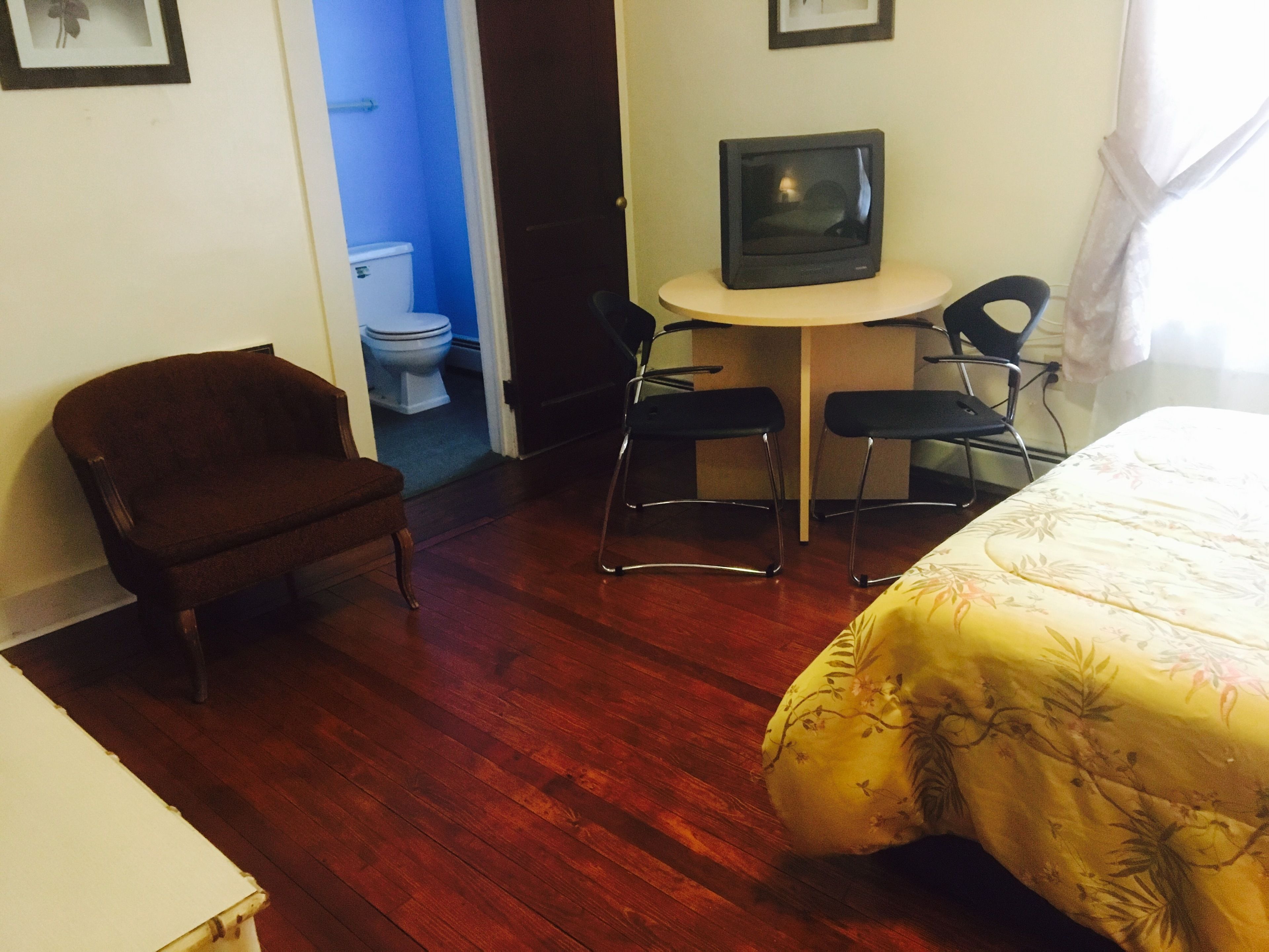 Ett hotellrum med trägolv, en säng med ett blommigt överkast, en brun fåtölj, ett litet bord med en tv och en stol.