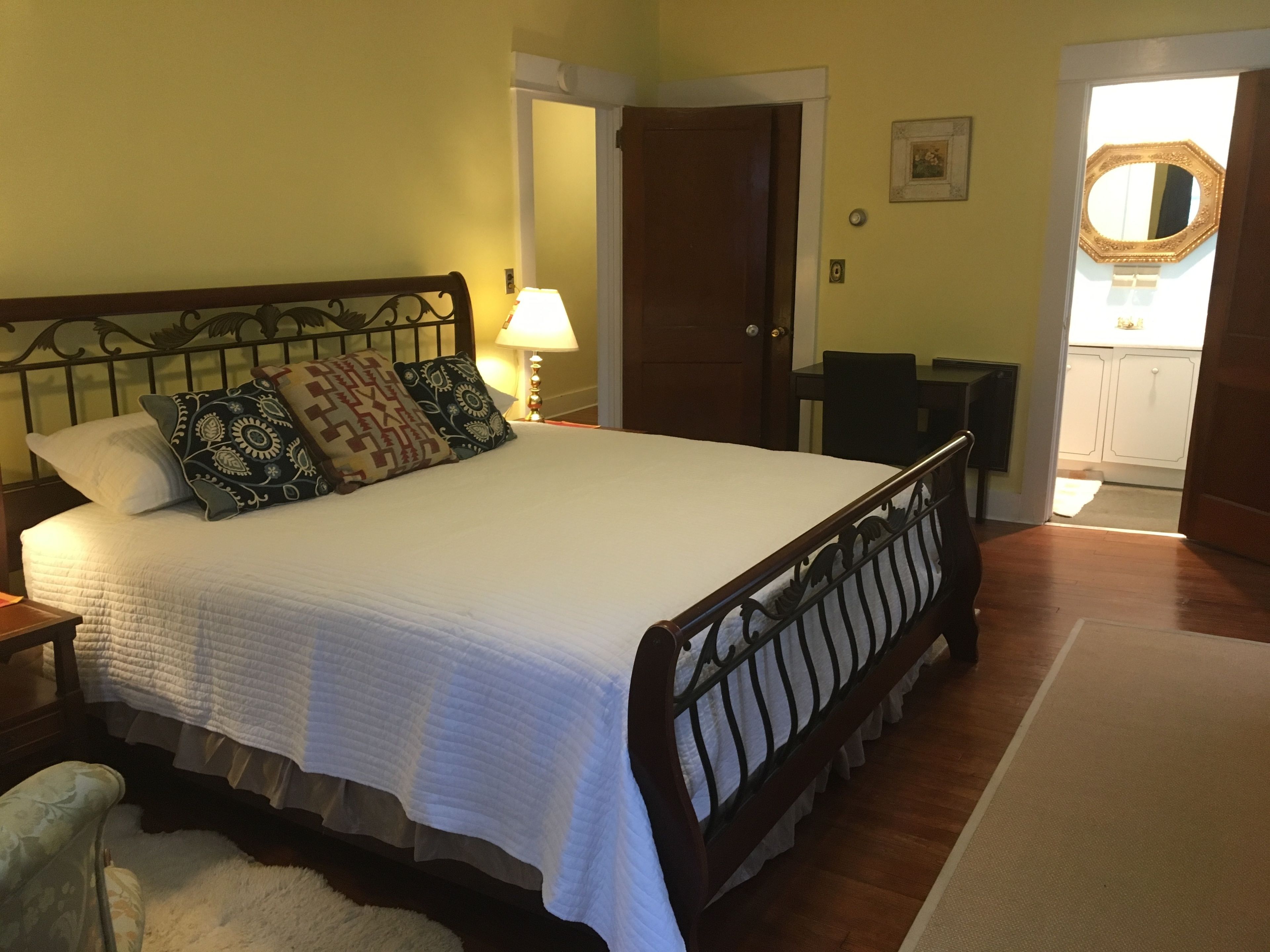 Una cama bien hecha con cabecera de madera y varios almohadones, una mesita de noche con lámpara, y vista a una cocina a través de una puerta abierta.