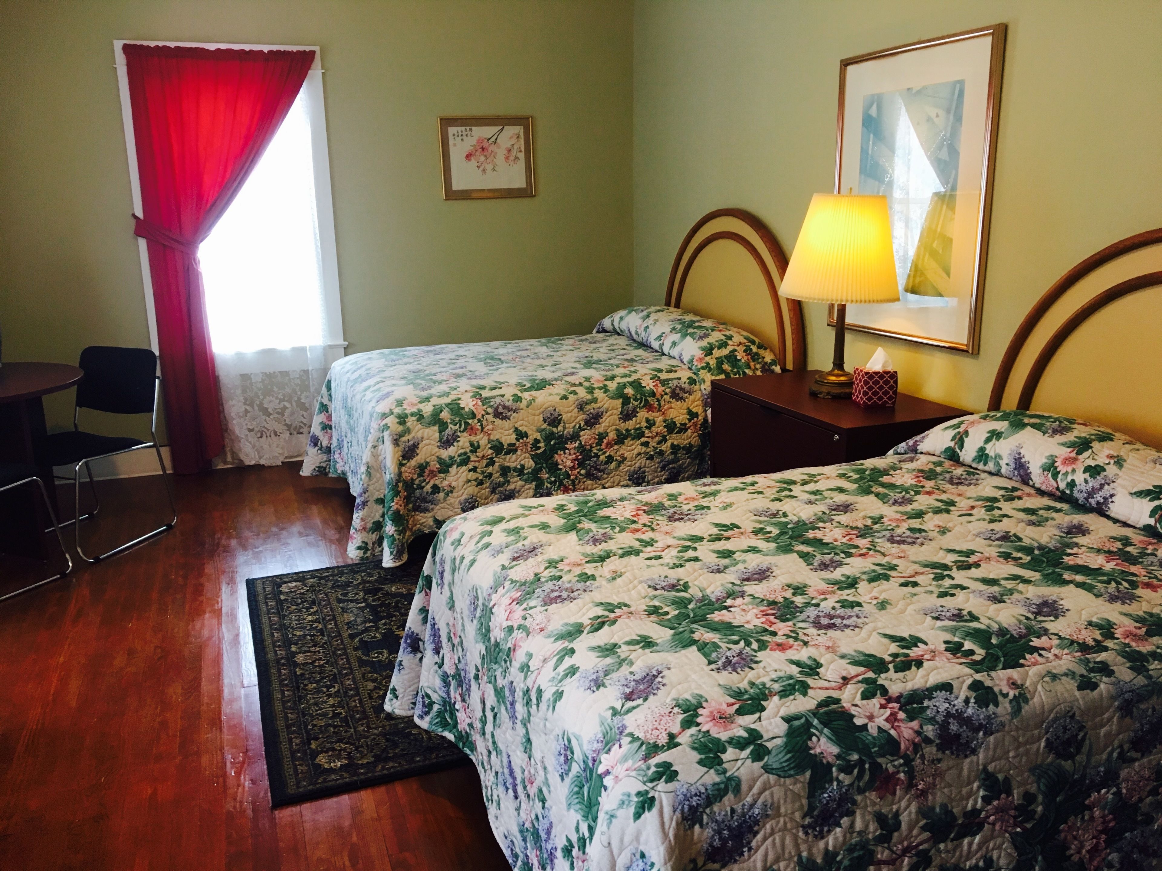 Une chambre avec deux lits, une petite table, une chaise, une lampe et une fenêtre avec des rideaux rouges.