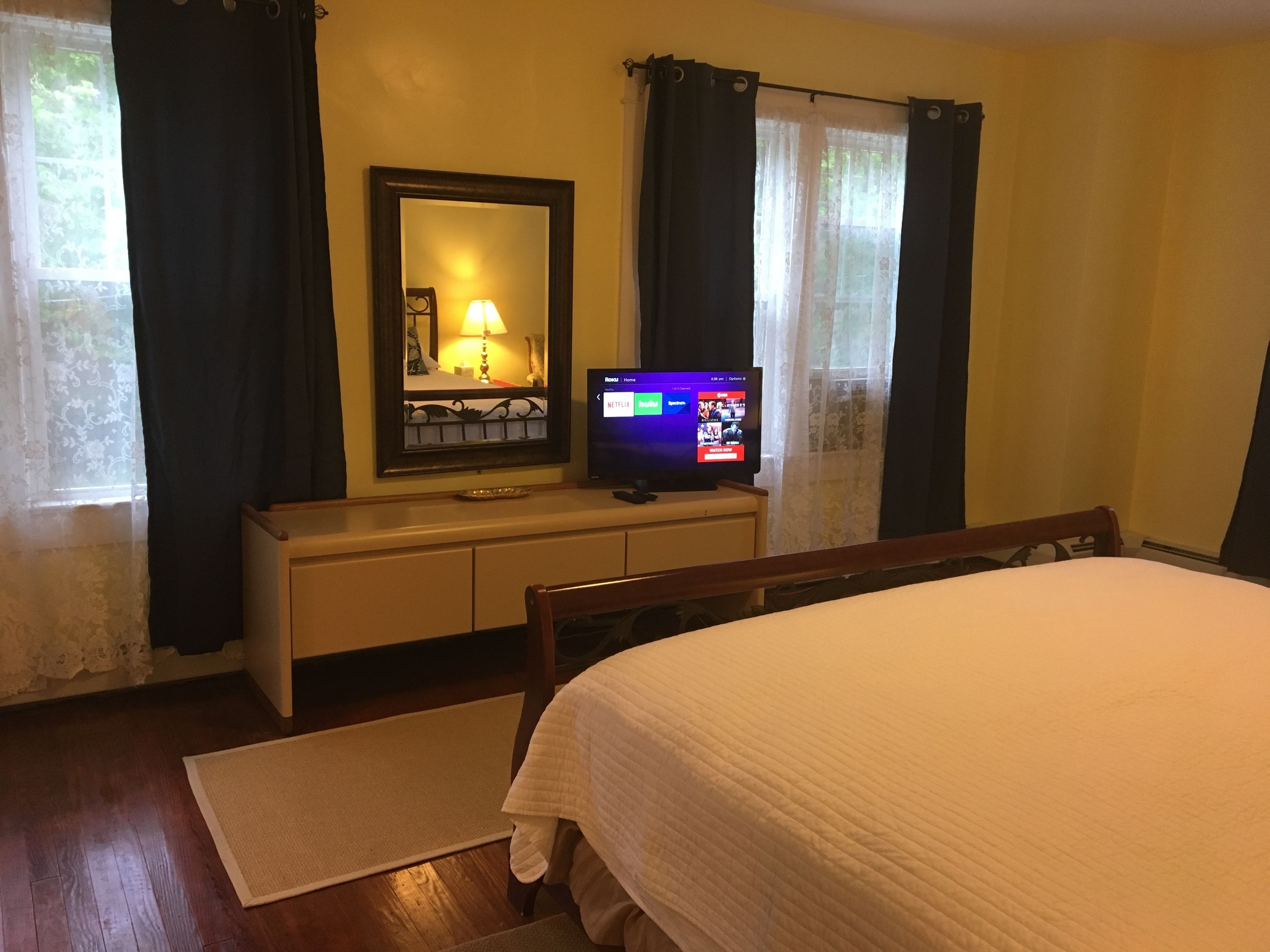 A hotel room with a bed, a TV on a stand, a dresser, a mirror, and a window with curtains.