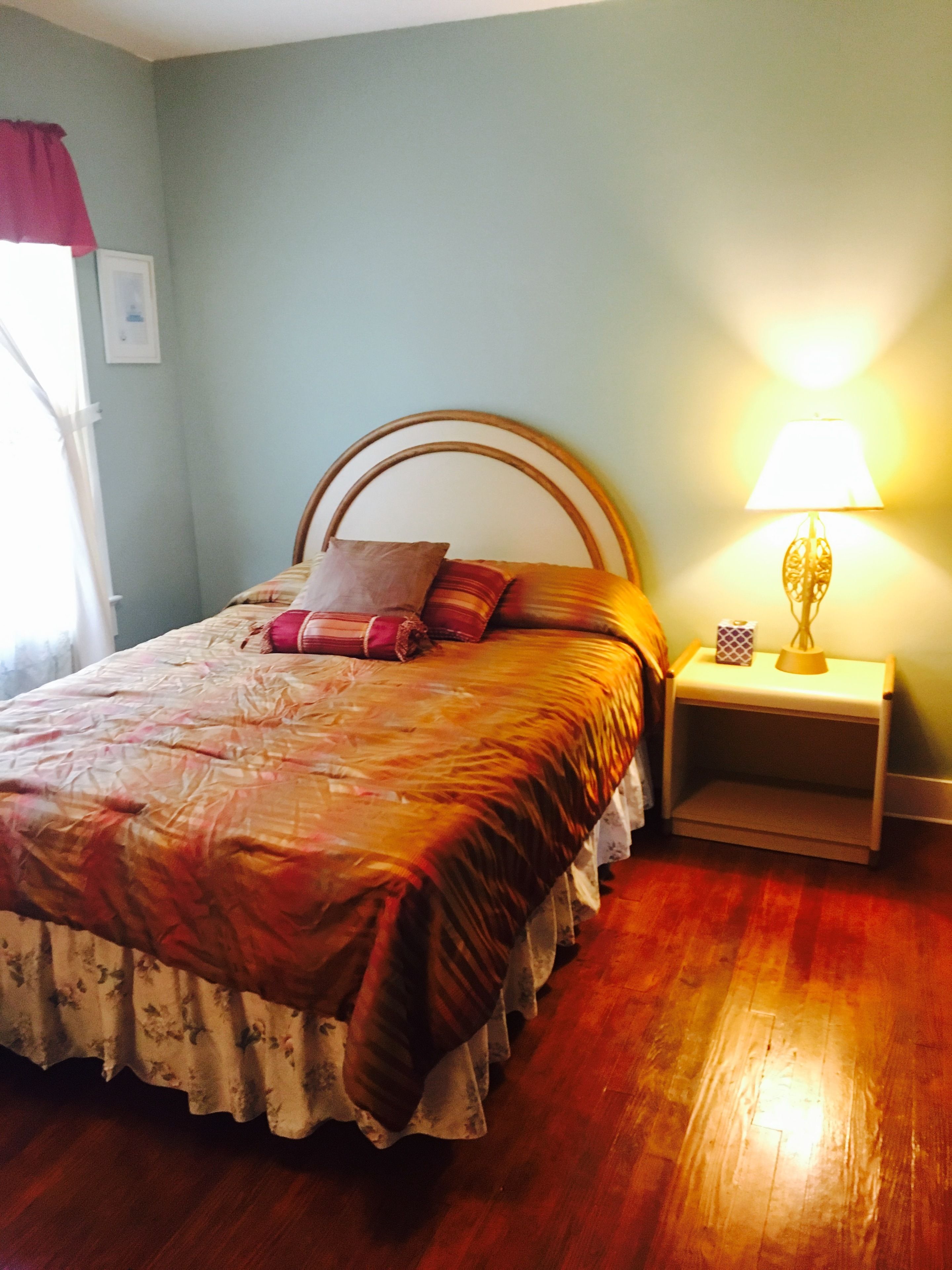 A neatly made bed with a floral bedspread and multiple pillows, a bedside table with a lamp, and a window with a red curtain.