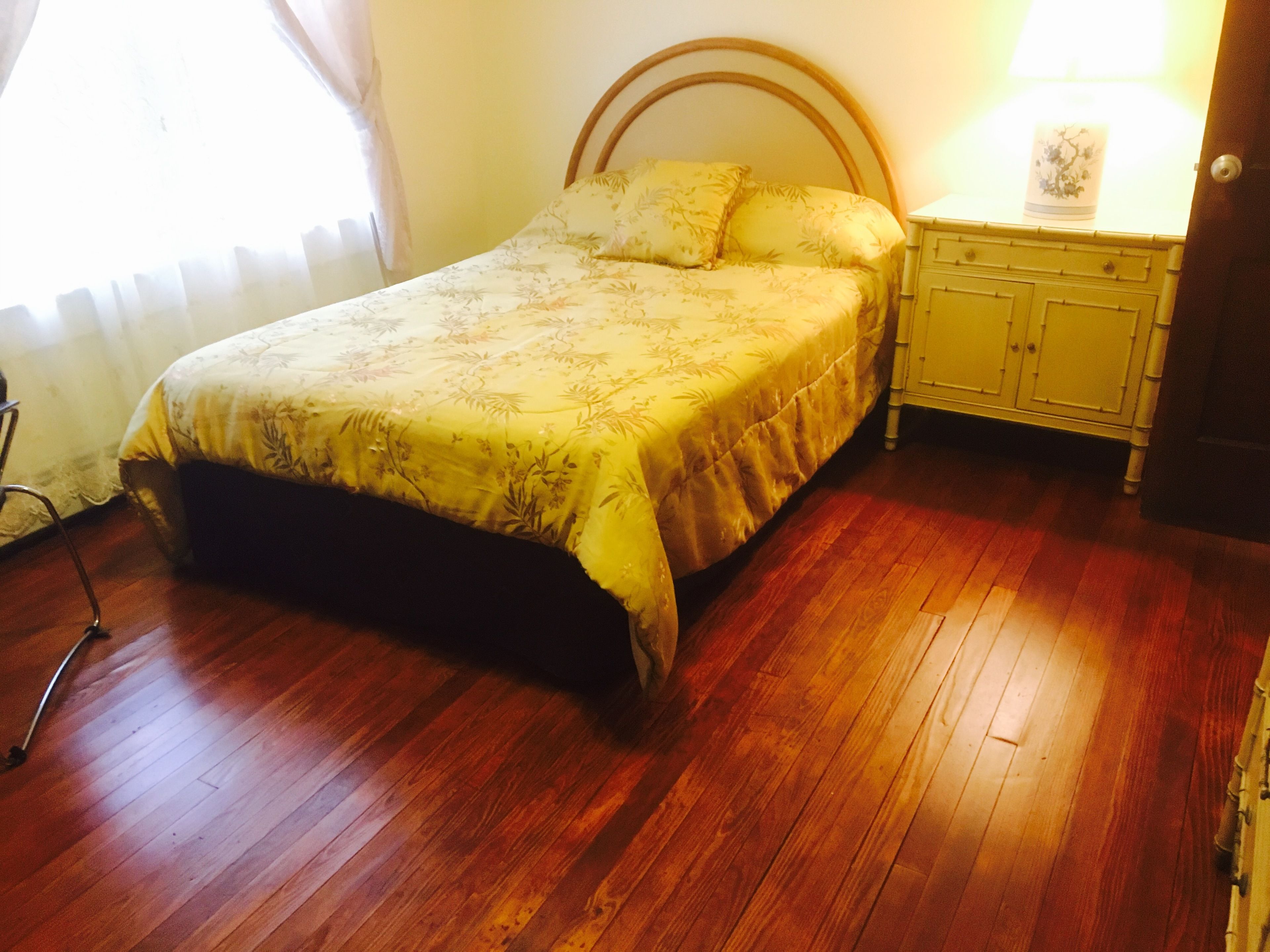Un dormitorio con cama, suelo de madera, mesita de noche, lámpara y ventana con cortinas.