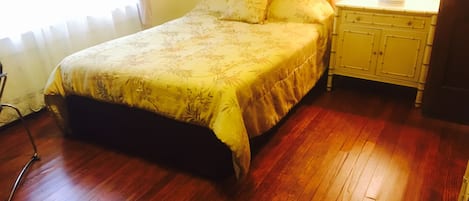Une chambre à coucher avec un lit, un plancher en bois, une table de chevet, une lampe et une fenêtre avec des rideaux.