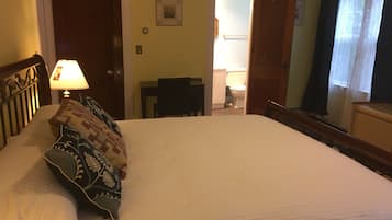 En snyggt bäddad säng med mönstrade kuddar, en sänglampa och en trägavel. I hörnet står ett litet bord och en stol, och genom en öppen dörr kan man se in i badrummet.