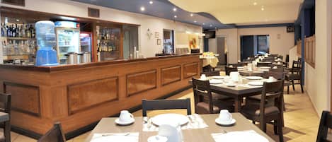 Un restaurante con paneles de madera, una zona de bar con dispensador de agua y mesas dispuestas con vajilla blanca.