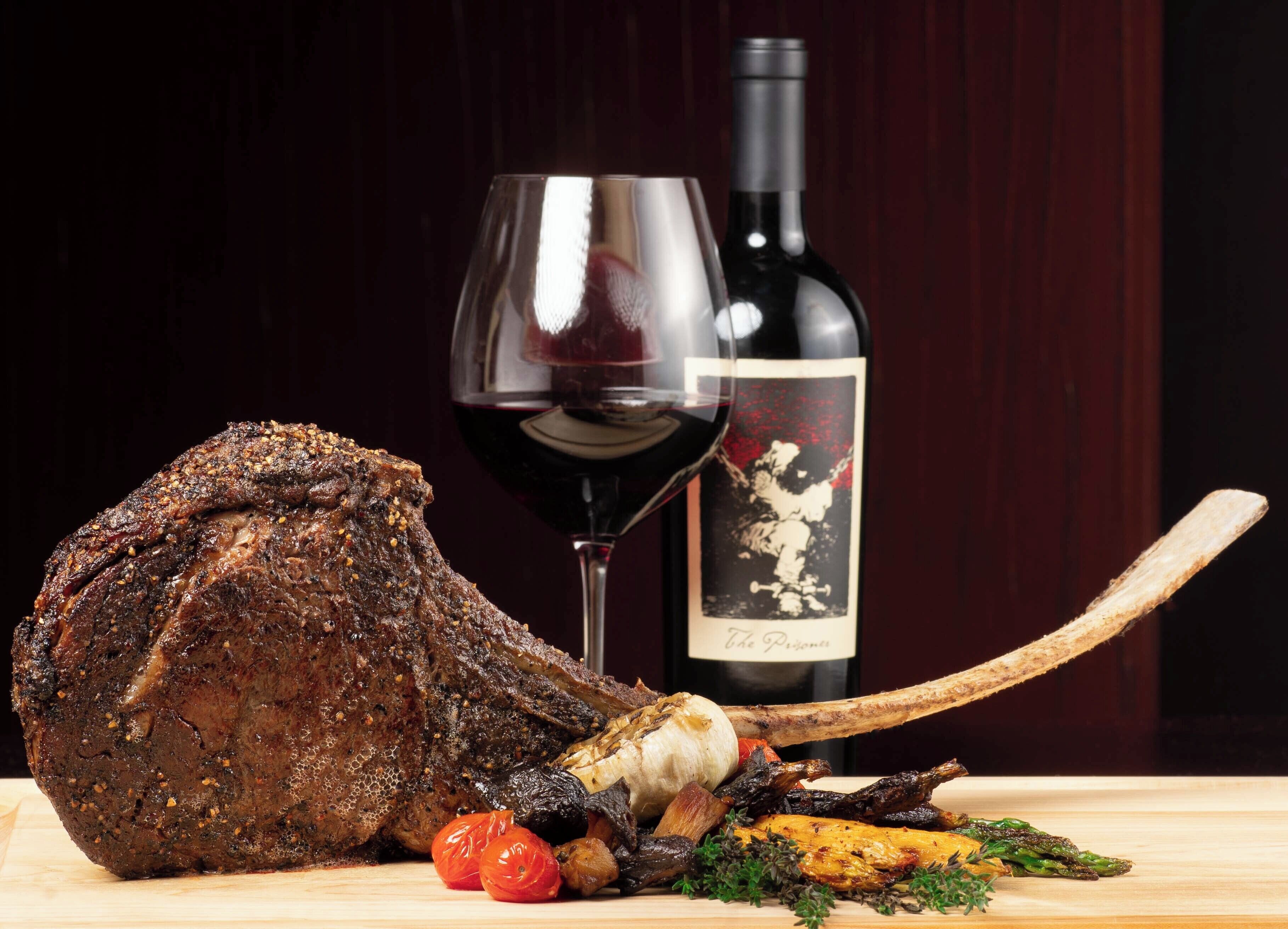 Um pedaço de pernil assado com legumes e uma garrafa de vinho sobre uma tábua de madeira.