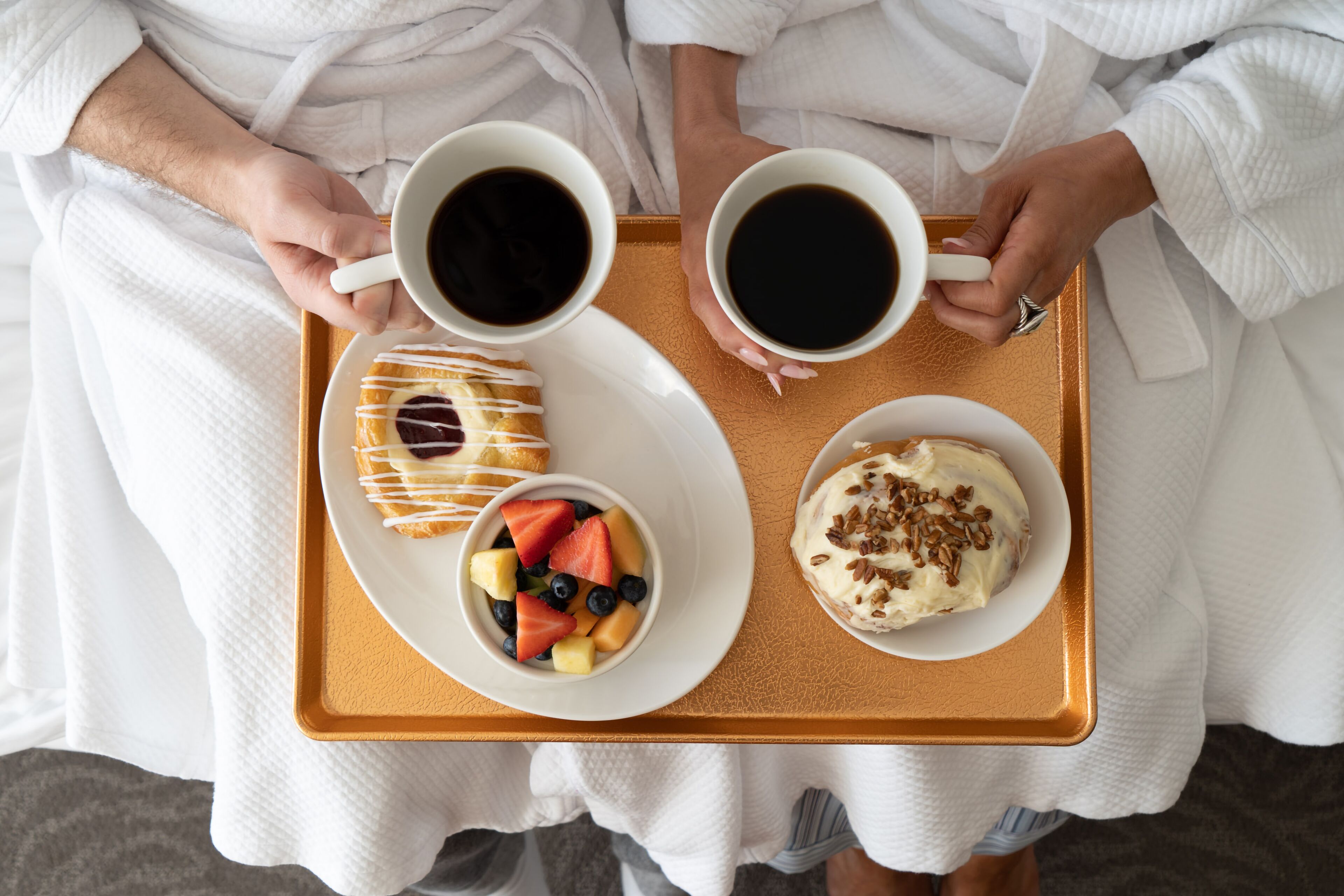 Una bandeja con dos tazas de café, un pastelito con glaseado blanco y un bol de fruta con fresas, arándanos y melón.