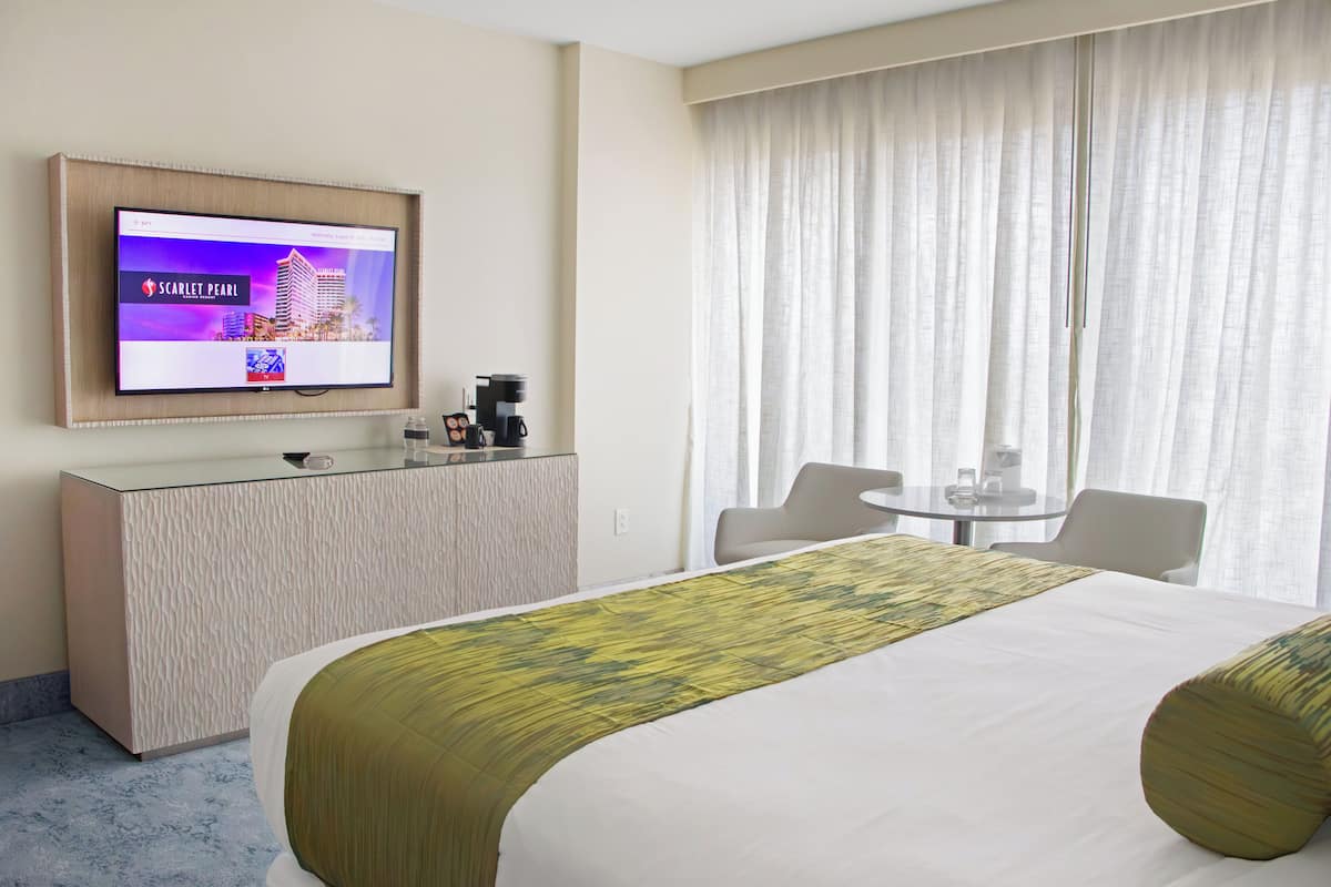 Ein Hotelzimmer mit einem Bett, einem an der Wand befestigten Fernseher, einem kleinen Tisch mit Stühlen und einem Fenster mit Vorhängen.