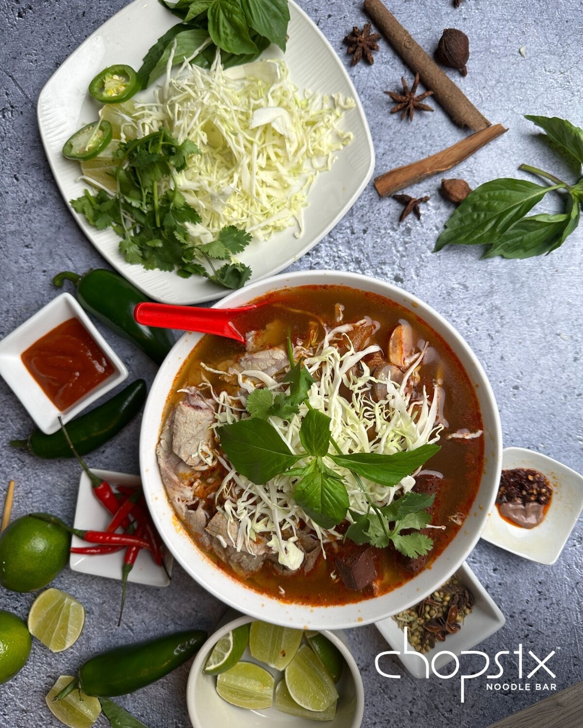A bowl of soup with various garnishes and side dishes, including sliced jalapeños, cilantro, and lime wedges.