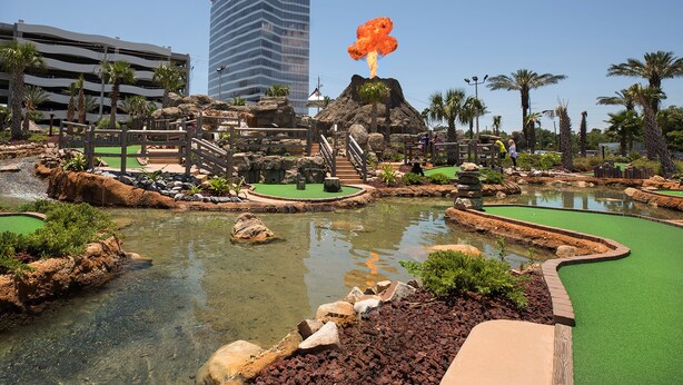 Un parcours de minigolf avec un obstacle sur le thème d’un volcan, des palmiers et, au fond, un parking.