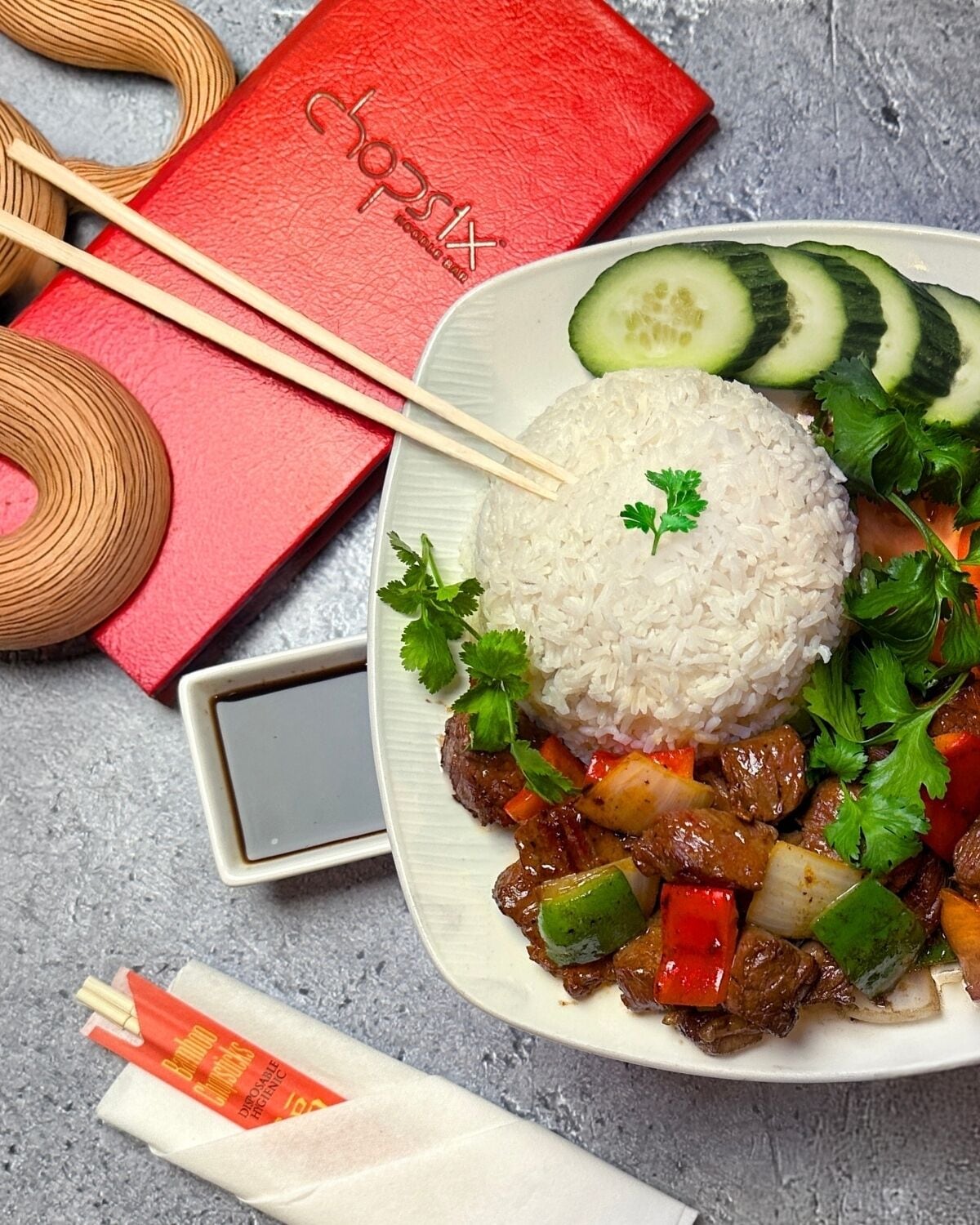 A plate of beef stir-fry with vegetables and rice, accompanied by chopsticks, a red box, and a small dish of sauce.