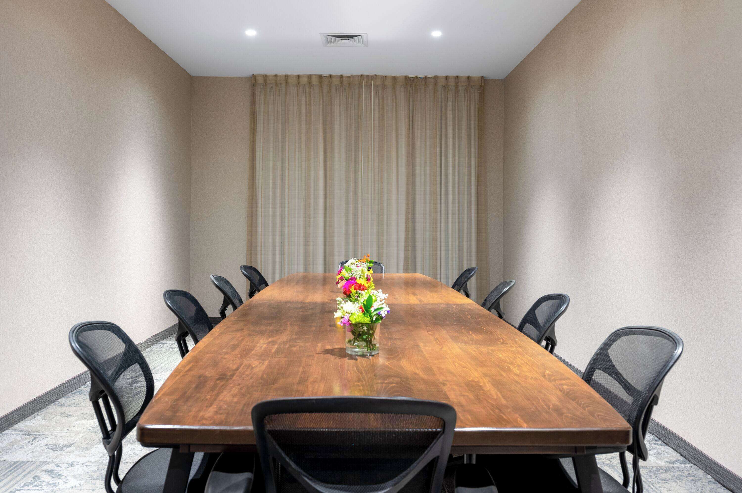 Una sala de conferencias con una larga mesa de madera, ocho sillas negras y un jarrón con flores.
