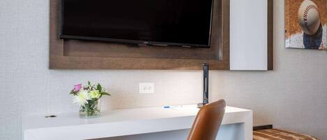 A modern hotel room with a flat-screen TV mounted on the wall, a white desk with a vase of flowers, a brown leather chair, and a framed baseball picture.