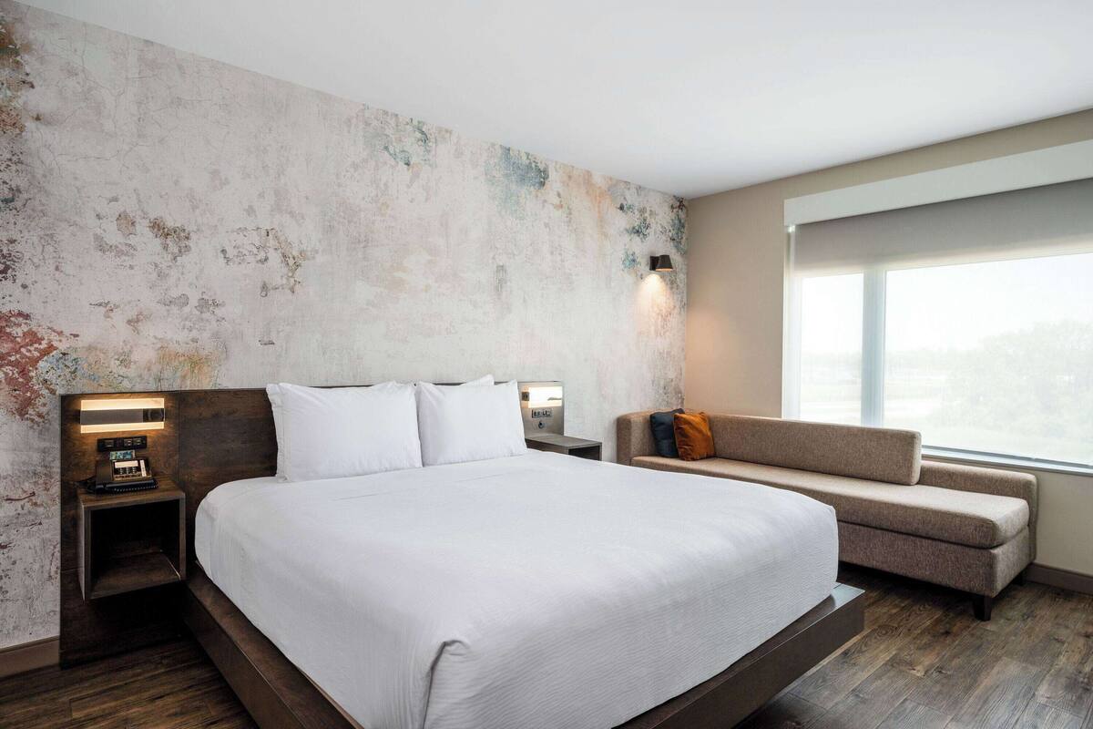 Une chambre d’hôtel moderne dotée d’un grand lit, d’une table de chevet avec une lampe, d’un canapé et d’une grande fenêtre donnant sur la ville.