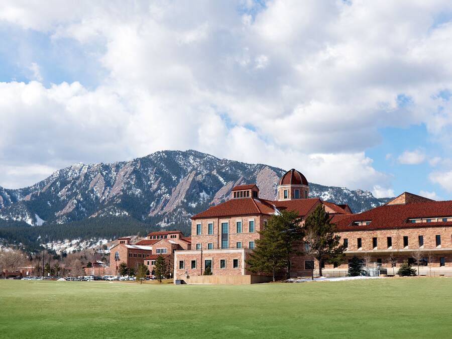 A large building with a central dome and multiple wings, surrounded by a green field and mountains in the background.