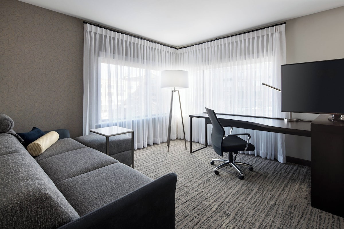 A hotel room with a grey sofa, a desk with a chair, a television, and a floor lamp.