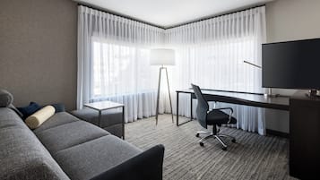 A hotel room with a grey sofa, a desk with a chair, a television, and a floor lamp.