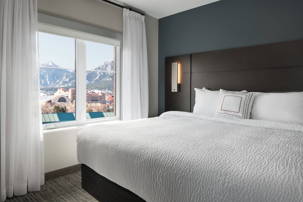 A hotel room with a large bed, a view of mountains, and a cityscape through the window.